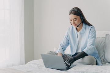 Disabled girl in headset typing on laptop using bionic prosthetic arm, sitting on bed at home