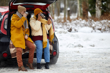 Full length portrait of young couple sitting in car trunk in winter forest while enjoying hot cocoa, copy space