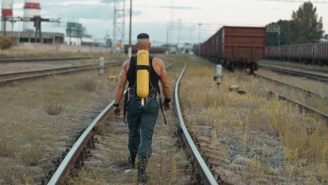 A young muscular man with drager on his back goes along the railroad tracks at marshalling yard next to the plant. Back view