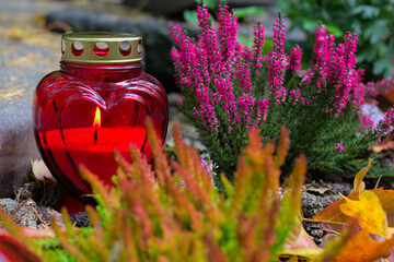 A red heart-shaped candle on a grave  in a cemetery on an autumn day. All Saints Day. Copy space, shallow depth of field. - obrazy, fototapety, plakaty