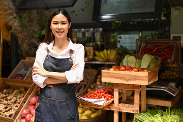 Portrait of friendly woman organic vegetable stall owner in apron standing with crossed arms and smiling at camera