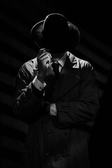 A dark silhouette of a man in a coat and hat in the noir style. A dramatic portrait in the style of detective films of the 1950s and 60s. The silhouette of a spy. - obrazy, fototapety, plakaty