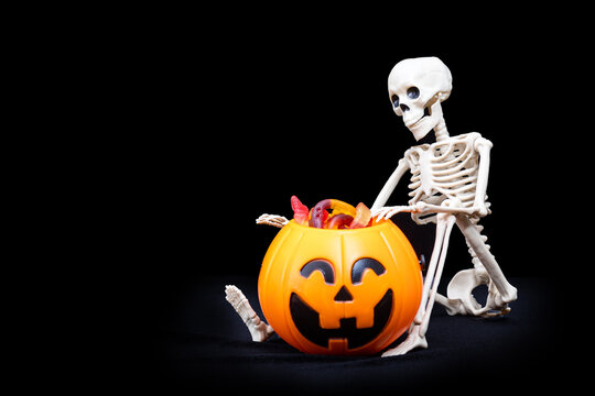 A Seated Skeleton Pulls Out Of A Bucket In The Shape Of A Pumpkin Candy On A Black Background, Copy Space. Halloween Trick Or Treat