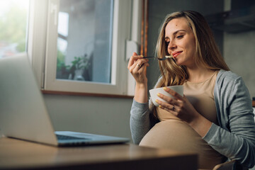 pregnant woman eating fruit salad and using laptop at home