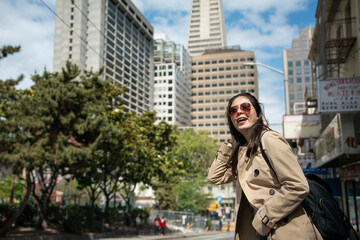 Fototapeta premium cheerful asian Korean female traveler wearing sunglasses running fingers through hair and looking at nice city landscape while waiting for bus at roadside in san Franciscoâs Chinatown