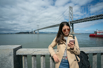 stylish asian female tourist leaning against outdoor railing and reading online guide on phone at leisure under blue sky with Oakland bay bridge at background at san Francisco bay