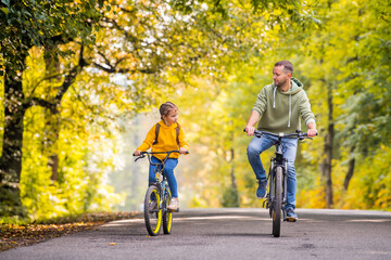 Happy father and daughter ride bicycles in autumn park on sunny day.