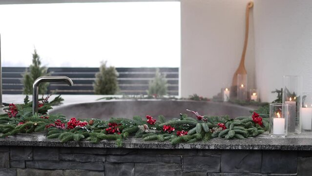 Steaming cast iron hot tub with spa in winter with snow and decorated candles. Christmas twigs with guelder rose and pine cones. 4K slow motion video
