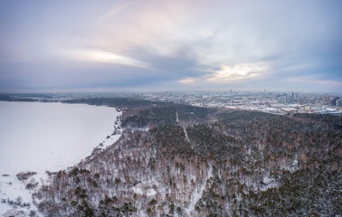 Fototapeta na wymiar Snow-covered forest on lake shore with ice at sunset and the city on horizon, auerial view
