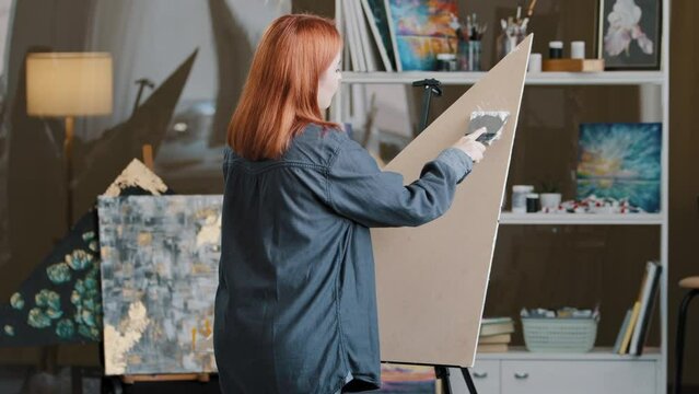 Red-haired Caucasian woman lady girl painter student of art school dancing indoor painting drawing creative masterpiece on canvas with white plaster spatula glue draw with music cheerful paint