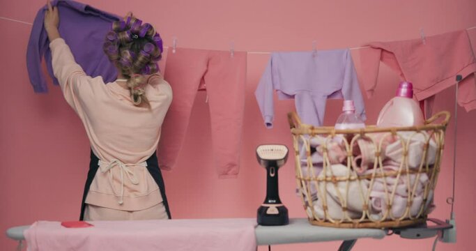 Woman with purple curlers hangs up washed children's clothes. Girl holds sweater, hangs it and pins it with clothespins. Wife looks into camera and smiles, basket with clothes is standing on the side.