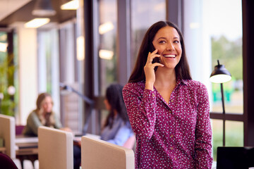 Businesswoman Standing By Partition In Open Plan Office Making Call On Mobile Phone