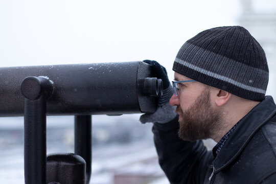 Close-up of bearded man in glasses looking through touristic scope, stationary observation binocle with coin-operated machine outdoors on winter landscape
