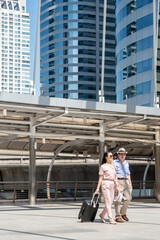Happy asian elderly senior couple of travelers walking in the big city dragging luggage suitcase on wheels. Holiday travel background.