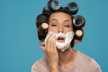 attractive young woman with hair curlers rollers applyes shaving foam on her face on a blue studio...