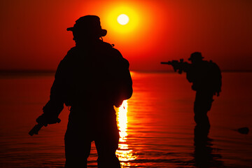 Army soldiers with rifles orange sunset silhouette in action during raid crossing river in the...