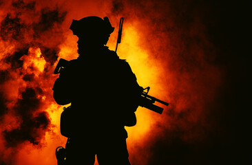 Silhouette of modern infantry soldier, elite army fighter in tactical ammunition and helmet, standing with assault service rifle in hands in the fire and smoke
