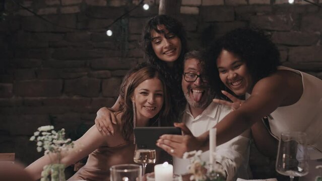 Cheerful young lesbian newlyweds having fun at wedding party in modern restaurant taking selfie on smartphone camera with parents