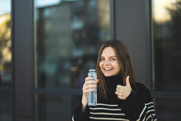 The girl holds a bottle of water in hands. Replenishment of the water balance. A girl drinks water from a reusable plastic bottle on the street. Water consumption.