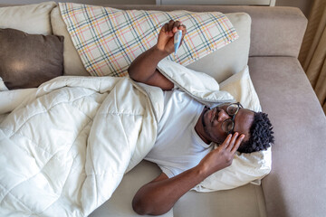 Young sick african guy with fever, checking body temperature with thermometer, lying on couch. Sick african american guy covered with scarf holding thermometer, having fever, empty space.
