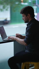Young man working remotely at co working office by window overlooking city street sidewalk....