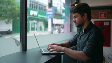 Man opening laptop seated at co working office by window overlooking city street sidewalk. Person works remotely