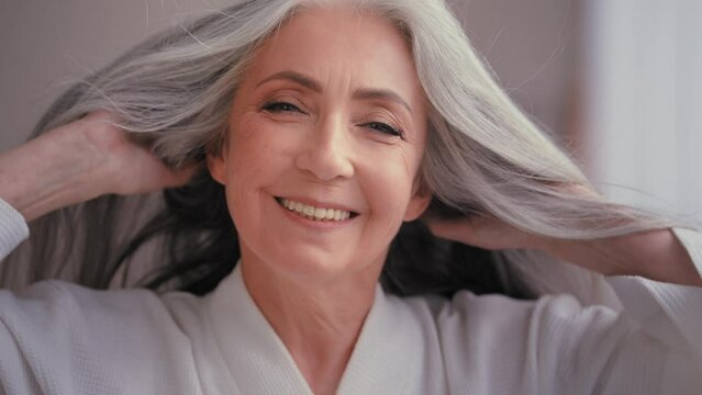 Gorgeous beautiful 60s old middle-aged mature Caucasian woman grandma senior older 50s lady smiling looking at camera at home pampering touching gray hair enjoy healthy hairstyle haircare procedure