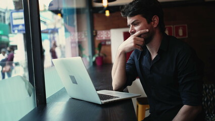 Concerned pensive young entrepreneur man in front of laptop computer seated by window at cafe co working remote workplace