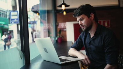 Concerned pensive young entrepreneur man in front of laptop computer seated by window at cafe co working remote workplace