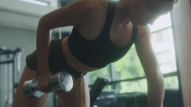 Close up tilt up shot fit woman in sports bra and shorts doing single arm dumbbell rows on bench while working out in gym