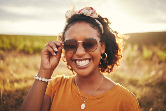 Face, fashion and nature with a black woman outdoor on a field with green grass in sunglasses and a smile. Happy, vacation and summer with a young female standing outside against a sky background