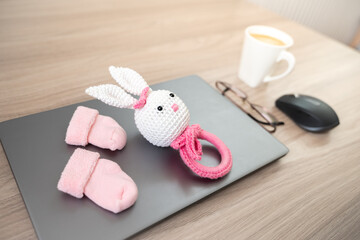 Working in a decree. Maternity leave concept. Closed laptop, pink toy and socks on a laptop, mouse, cup of coffee and glasses on a wooden table.