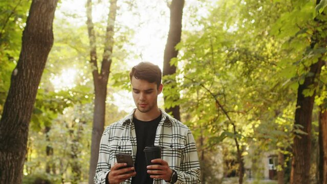 Tracking shot of a handsome man walking through green alley early in the morning, looking busily on his smartphone, using app on his way to work while sipping hot beverage out of a disposable cup