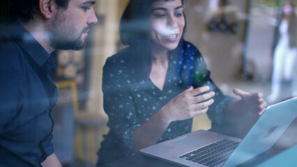 Young hispanic woman explaining work or study to male colleague seen through window reflection by city sidewalk street. Focused female leader pointing laptop screen at business workplace