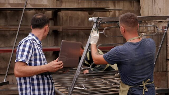 Two blacksmiths make metal railings for stairs, one grinds metal, the other measures the angle with a special tool.