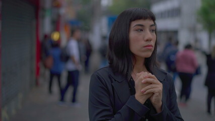 Stressed young hispanic woman standing in urban street fidgeting with hands. Preoccupied South American adult girl feeling nervous and preoccupation