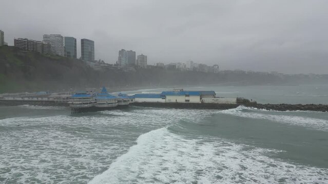 Aerial low angle dolly shot off the coast of costa verde in lima peru with the rosa nautica restaurant built on stones placed in the sea overlooking building on a cloudy morning