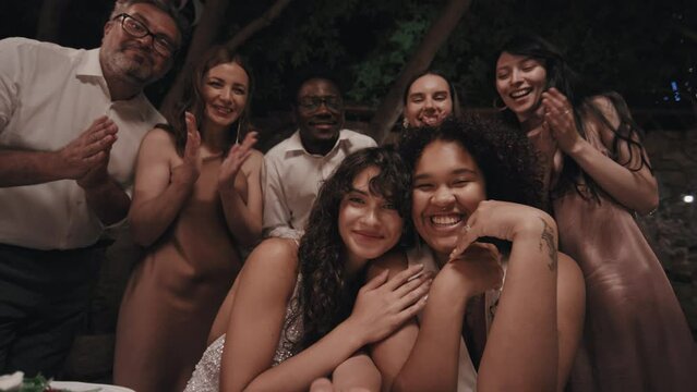 Low angle view of young lesbian couple demonstrating wedding rings on camera, their friends and relatives standing behind clapping hands