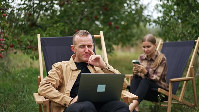 Positive mid aged man working remotely on his laptop. Lady sitting next in the garden chair looks at smartphone in her hands. Apple orchard background.