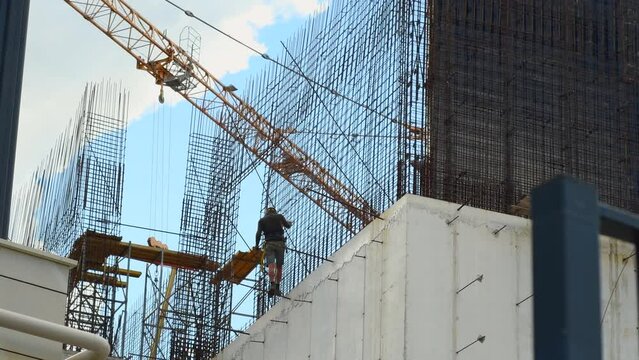 A construction worker in a white helmet walks along the unfinished wall of a building under construction. Reinforcement pins protrude from an unfilled concrete wall.