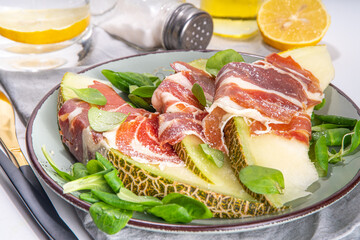 Fresh melon salad with prosciutto hamon and green leaves mix, autumn fruit vegetable salad, portion on plate, on white marble background copy space