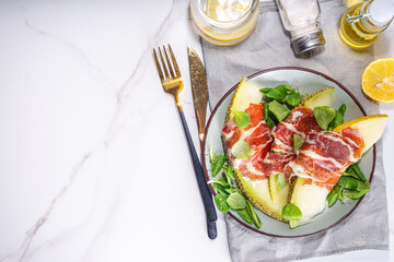 Fresh melon salad with prosciutto hamon and green leaves mix, autumn fruit vegetable salad, portion on plate, on white marble background copy space