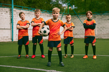 Teammates. Athletic boys in junior soccer team standing together at grass sport field. Football players in orange-black kits and boots.