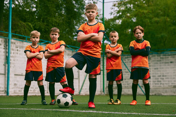 Teammates. Athletic boys in junior soccer team standing together at grass sport field. Football players in orange-black kits and boots. - Powered by Adobe