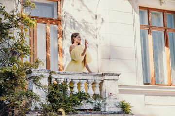 a beautiful smiling and kind woman in a gorgeous yellow dress stands on the balcony of an old vintage house
