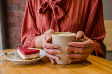 Young woman in red dress is sitting at table in cafe, red cake on saucer near, she is holding cup with drink in hands