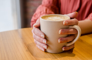 Young female hands holding and hugging a cup of coffee with latte art at wooden table in a cafe. Woman in red dress