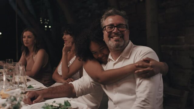 Happy Black lesbian bride sitting at festive table in restaurant in evening hugging her father-in-law smiling at camera