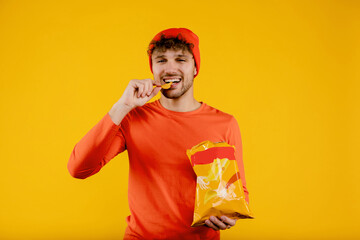 Young happy man holds puts chip in his mouth and holds pack of chips in another, isolated yellow...
