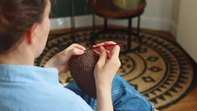 Back side young woman crocheting brown bag using pink hook and raffia yarn sitting at cozy home. Earning money on your handmade hobby concept.Knitting is tranquil method of calming the nervous system